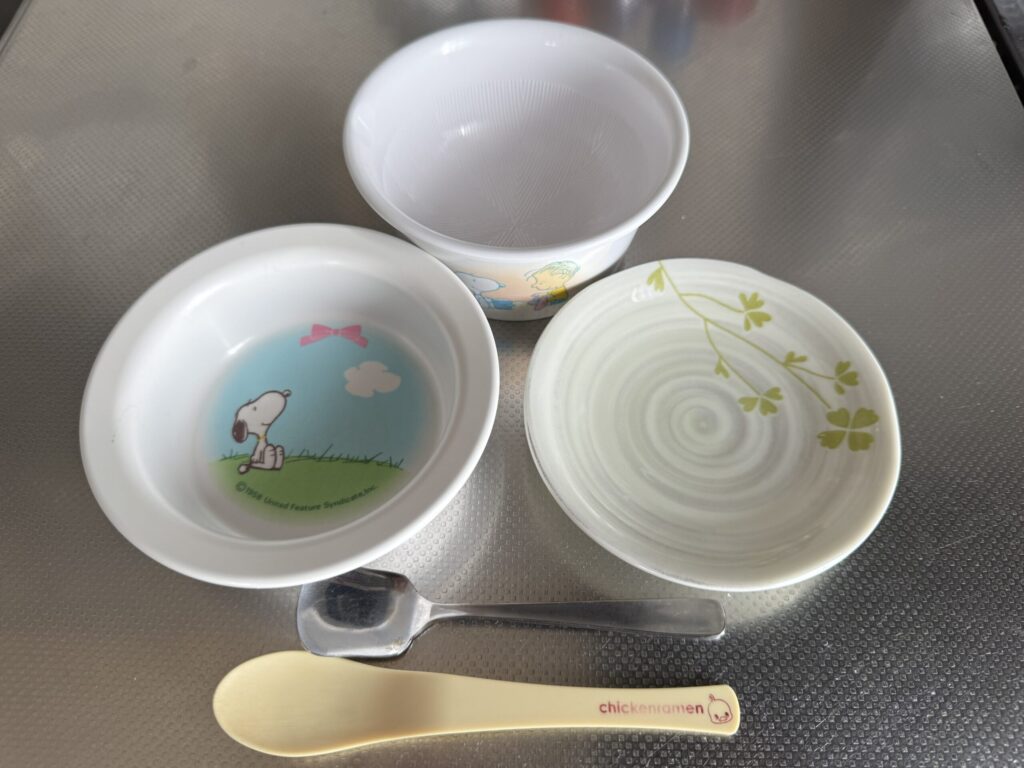 離乳食で実際に使っている道具。スヌーピー柄の子ども用の皿と容器、クローバー柄の陶器の小皿、金属のティースプーン、ひよこ柄のプラスチックスプーン