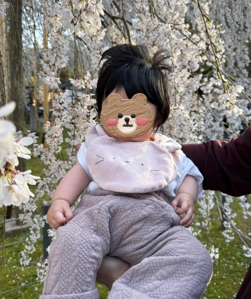 桜を背景に、ねこのスタイとピンクのキルトパンツ姿の生後6ヶ月の赤ちゃん